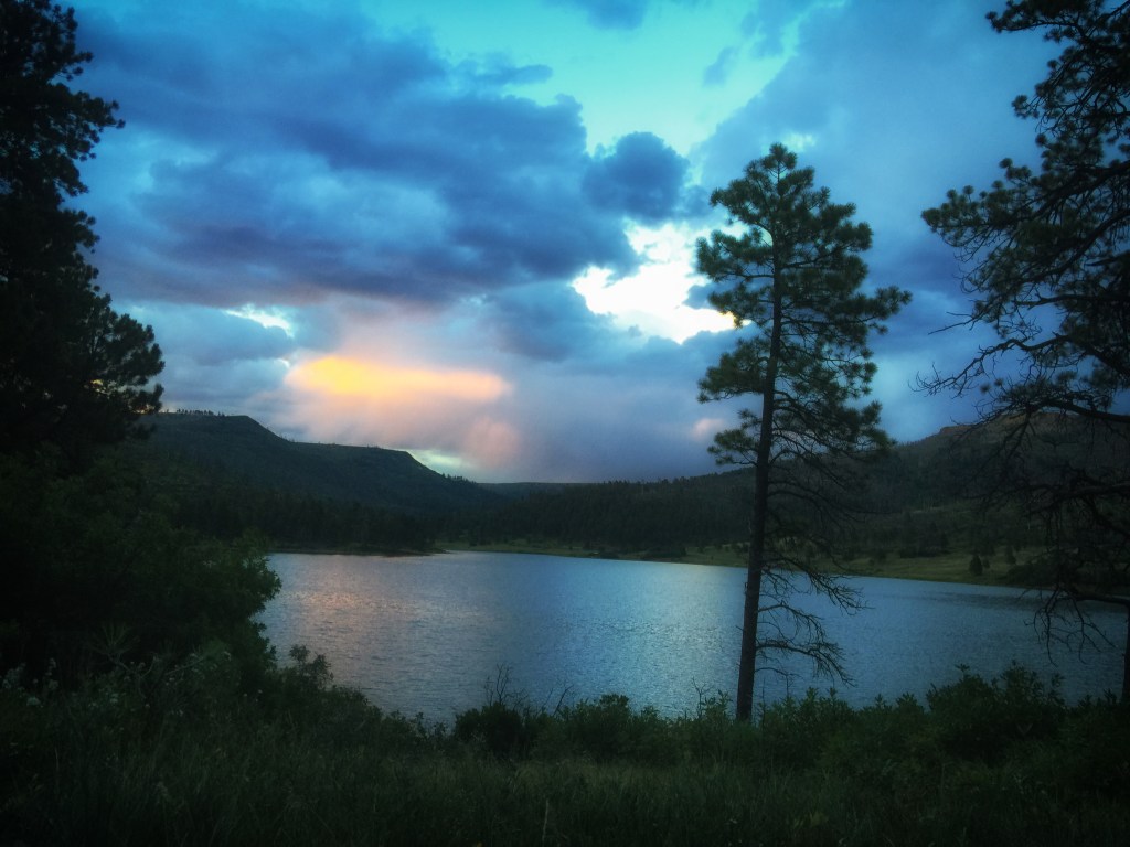 Lake Maloya under a dramatic cloudy sky with a break of warm light reflecting on the water, framed by pine trees